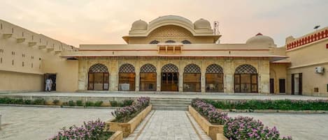 A courtyard with a central building featuring arched doorways and a dome, surrounded by flower beds and a tiled walkway.