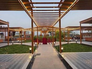 Un paseo techado al aire libre con vigas de madera y luces, que conduce a un área central con plantas en macetas y asientos.