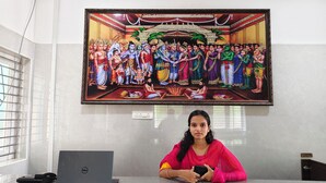 A person seated at a desk with a laptop and a smartphone, in front of a large framed painting depicting a traditional scene. - AA Residency (Tirupati)