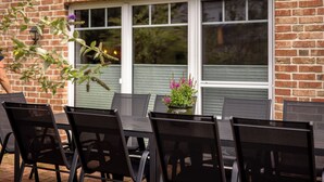 Une terrasse extérieure avec un mur en briques, une fenêtre avec des stores et une table entourée de chaises.