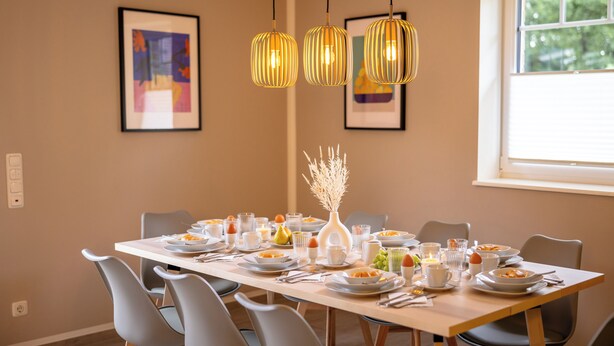 Un espace repas avec une longue table dressée pour un repas, trois lampes suspendues, des œuvres d’art encadrées accrochées aux murs et une fenêtre laissant entrer la lumière naturelle.