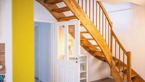 Un escalier en bois mène à une chambre située en mezzanine, avec une porte blanche et un meuble dont les portes sont en verre.