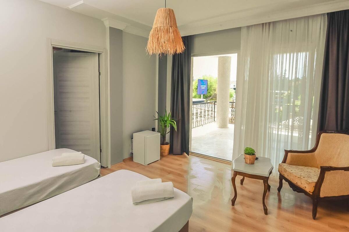 A hotel room with two beds, a small table, a chair, a hanging light fixture, and a view of the outside.