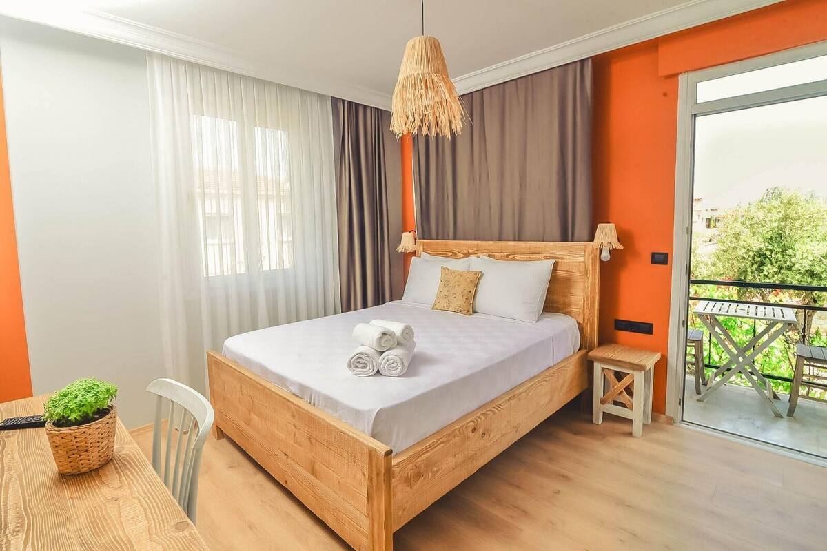 A wooden bed with white linens, a small bedside table, a hanging lamp, and a view of greenery through a sliding glass door.