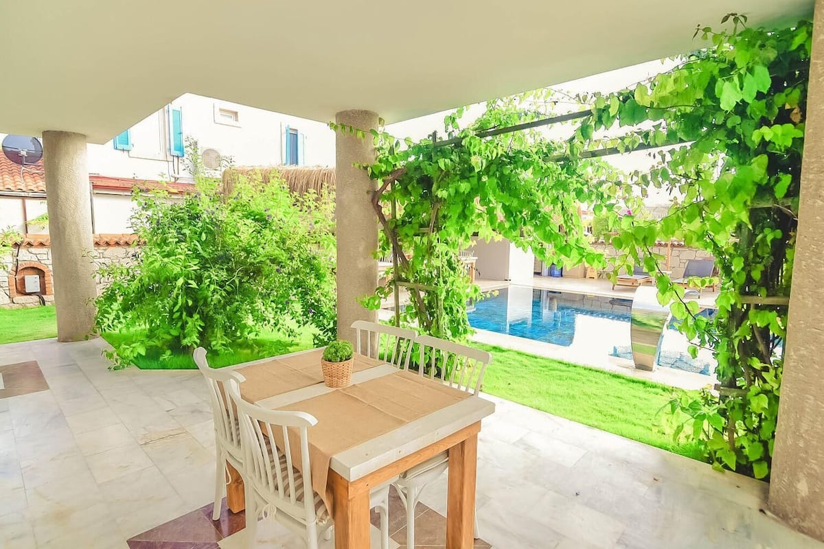 A covered patio with a table and chairs, a pool, and greenery.