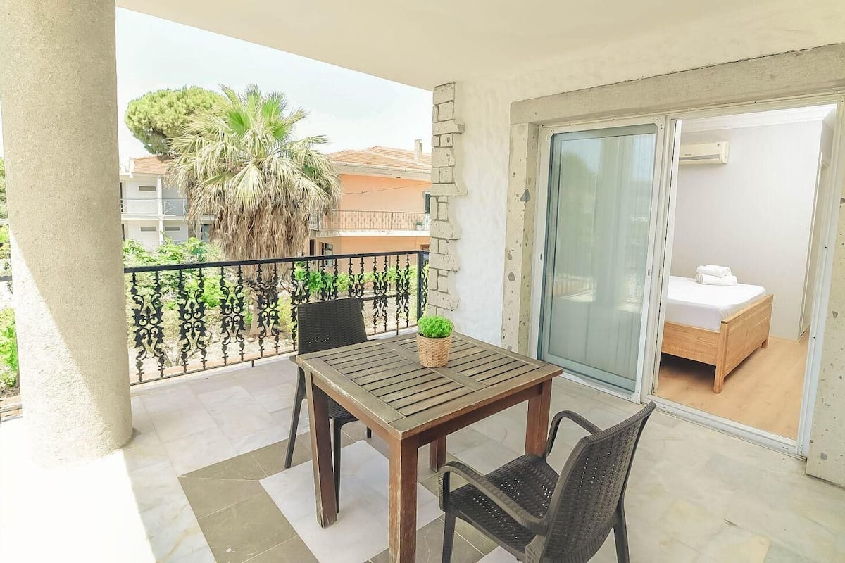 A balcony with a wooden table and chairs, a small potted plant, a palm tree, and a sliding glass door leading to a bedroom with a bed and a white towel.