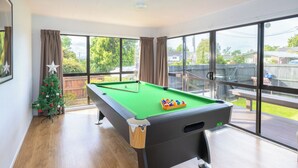 A pool table with a green felt surface, a Christmas tree, and a view of a backyard through large windows.