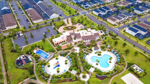 Vue aérienne d’un quartier résidentiel avec deux grandes piscines, un pont enjambant un plan d’eau et une végétation luxuriante tout autour.