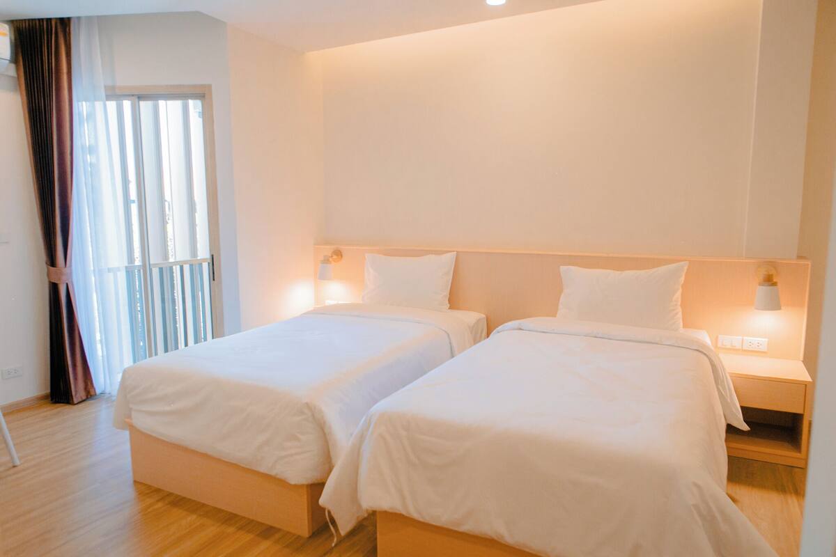 A hotel room with two single beds, a wooden floor, a large window with curtains, and bedside lamps.