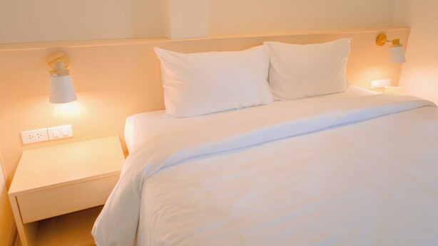 Una cama bien hecha con sábanas y almohadas blancas, una mesita de noche con lámpara, y una lámpara de pared.