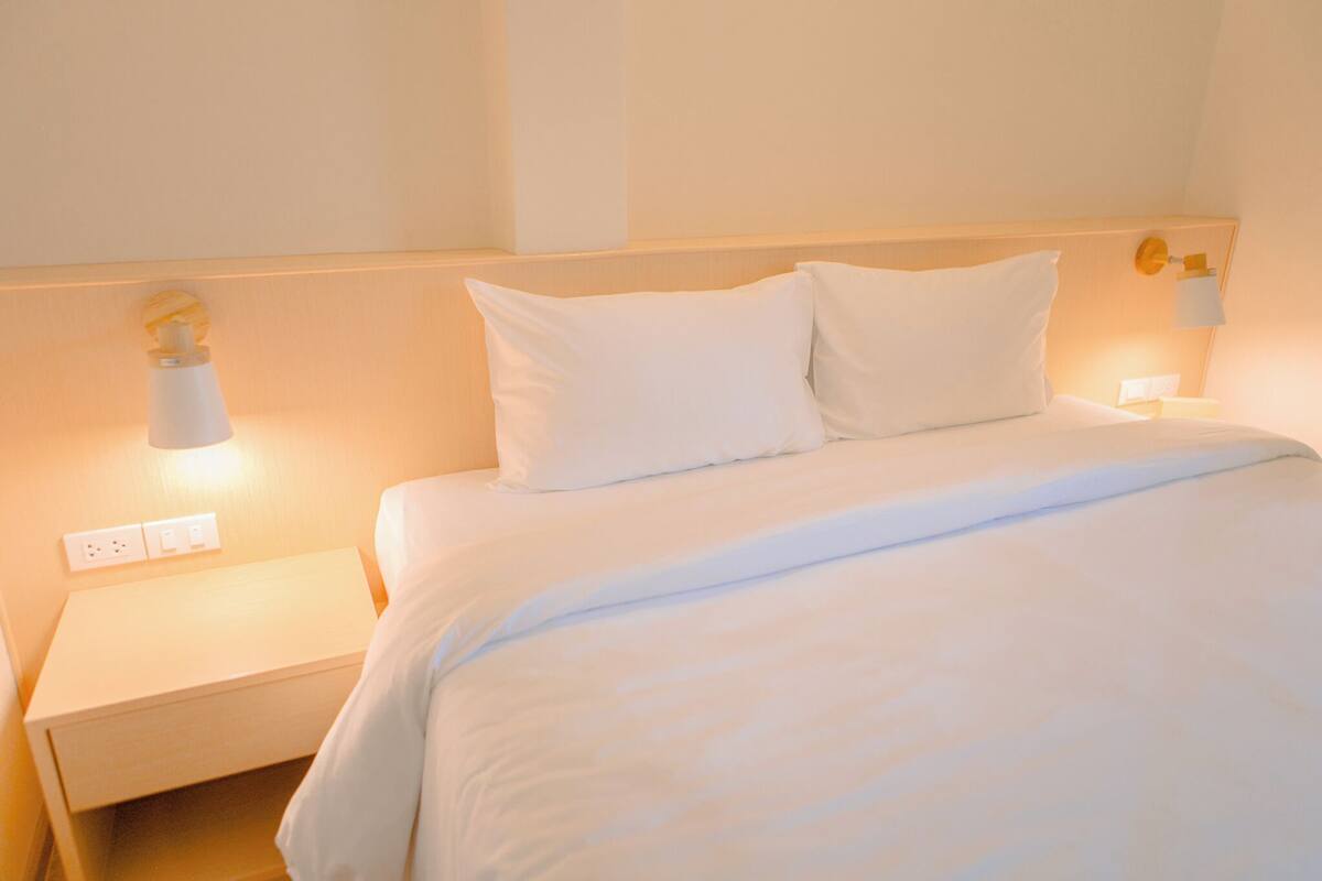 A neatly made bed with white linens and pillows, a bedside table with a lamp, and a wall-mounted light fixture.