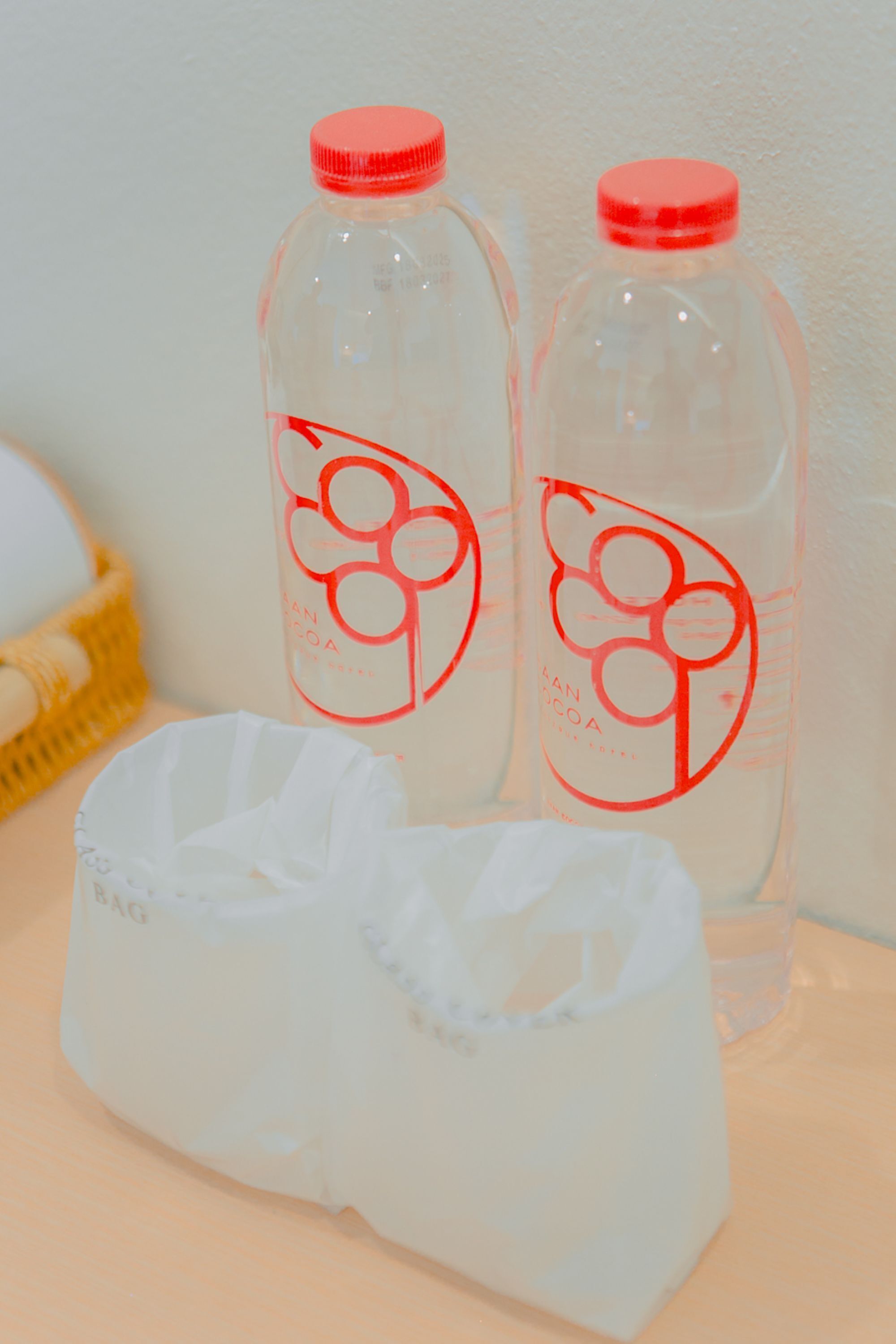 Two bottles with a red cap and a white label with a red logo, placed on a table next to a white plastic bag.