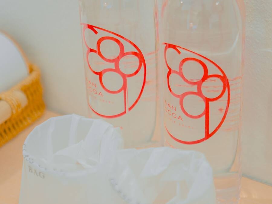 Two bottles with a red cap and a white label with a red logo, placed on a table next to a white plastic bag.