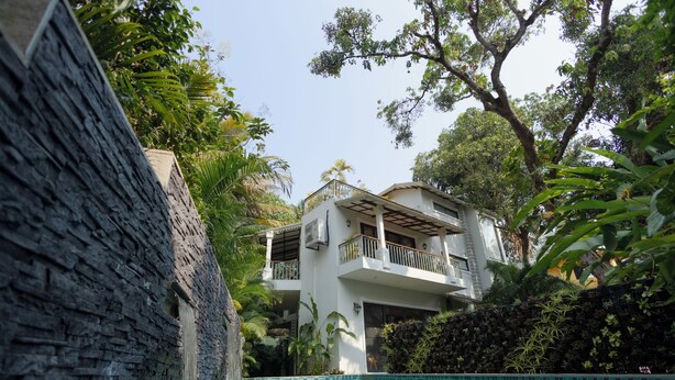 Un área de piscina con un muro de piedra, un edificio de dos pisos con balcones y abundante vegetación.
