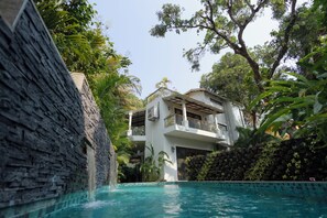 A pool area with a stone wall, a two-story building with balconies, and lush greenery. - Villa Anjuna By Akama Stays (Anjuna)