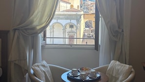 Una stanza con vista sulla città attraverso una finestra con tende bianche. Si nota un tavolino rotondo con un vassoio contenente tazze e piattini.