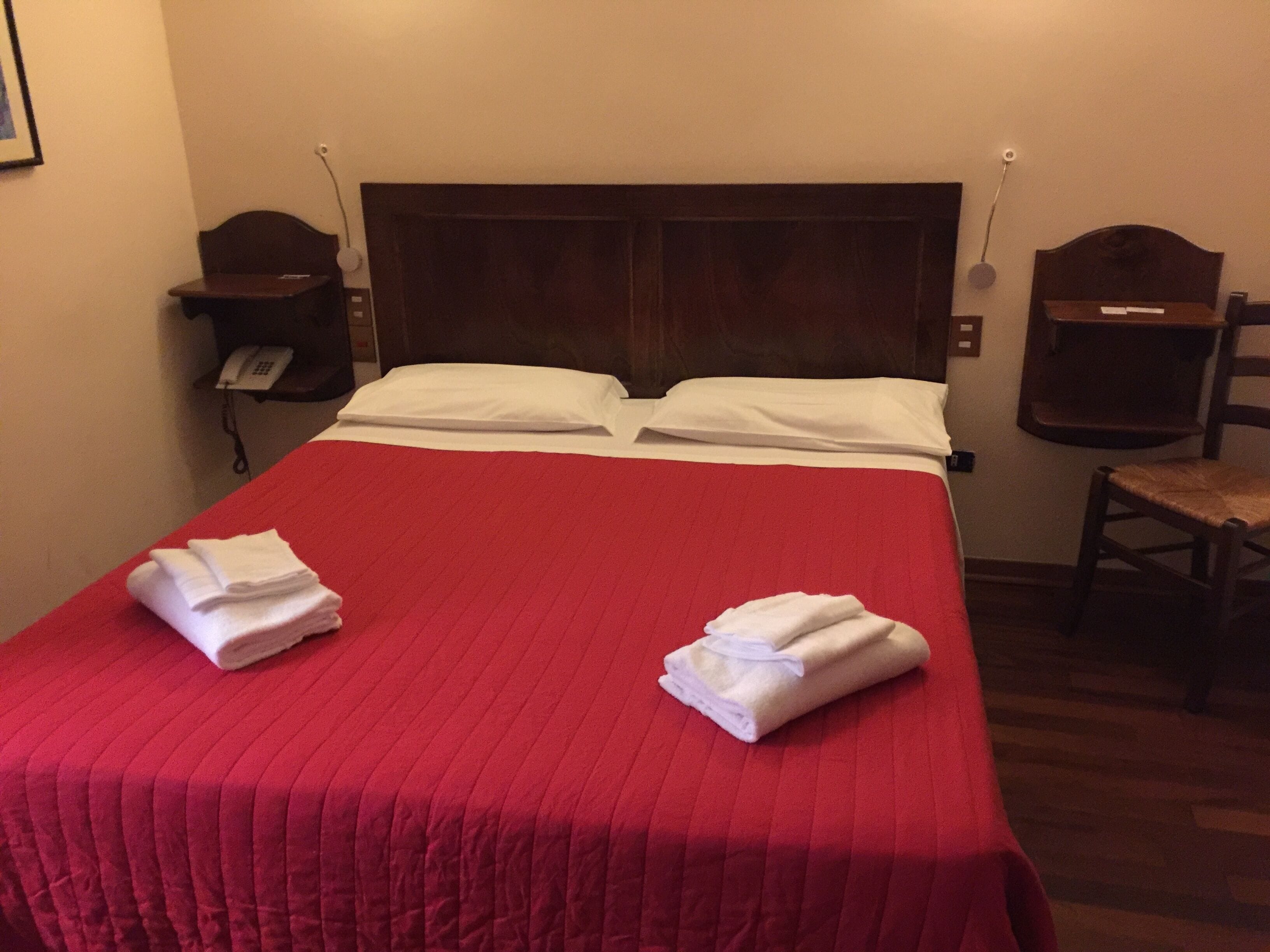 A hotel room with a red bedspread, two white pillows, a wooden headboard, a bedside telephone, and a wooden chair.