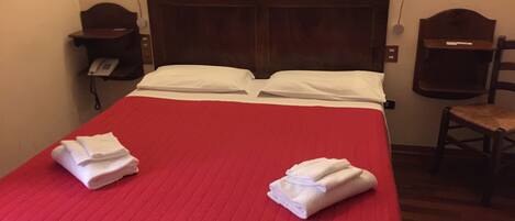 A hotel room with a red bedspread, two white pillows, a wooden headboard, a bedside telephone, and a wooden chair.