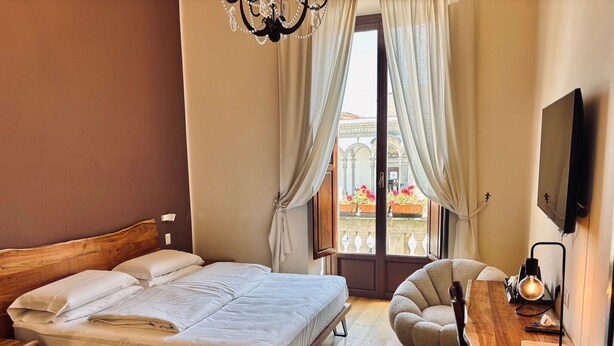 Una camera da letto con un letto di legno, una scrivania con una sedia, una TV e vista su un edificio attraverso una finestra con tende.