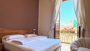 Una camera d'albergo con un letto grande, una sedia e un balcone affacciato su edifici e fiori.