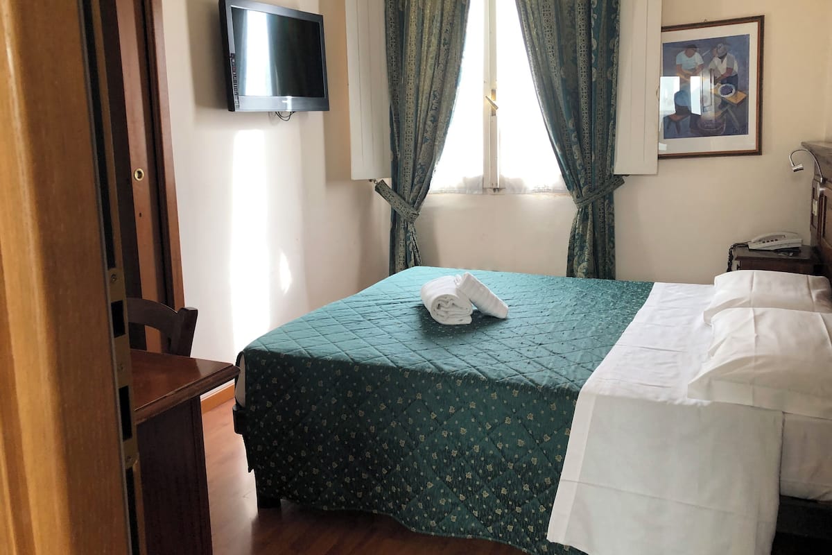 A hotel room with a bed, a TV mounted on the wall, a painting on the wall, and a window with curtains.