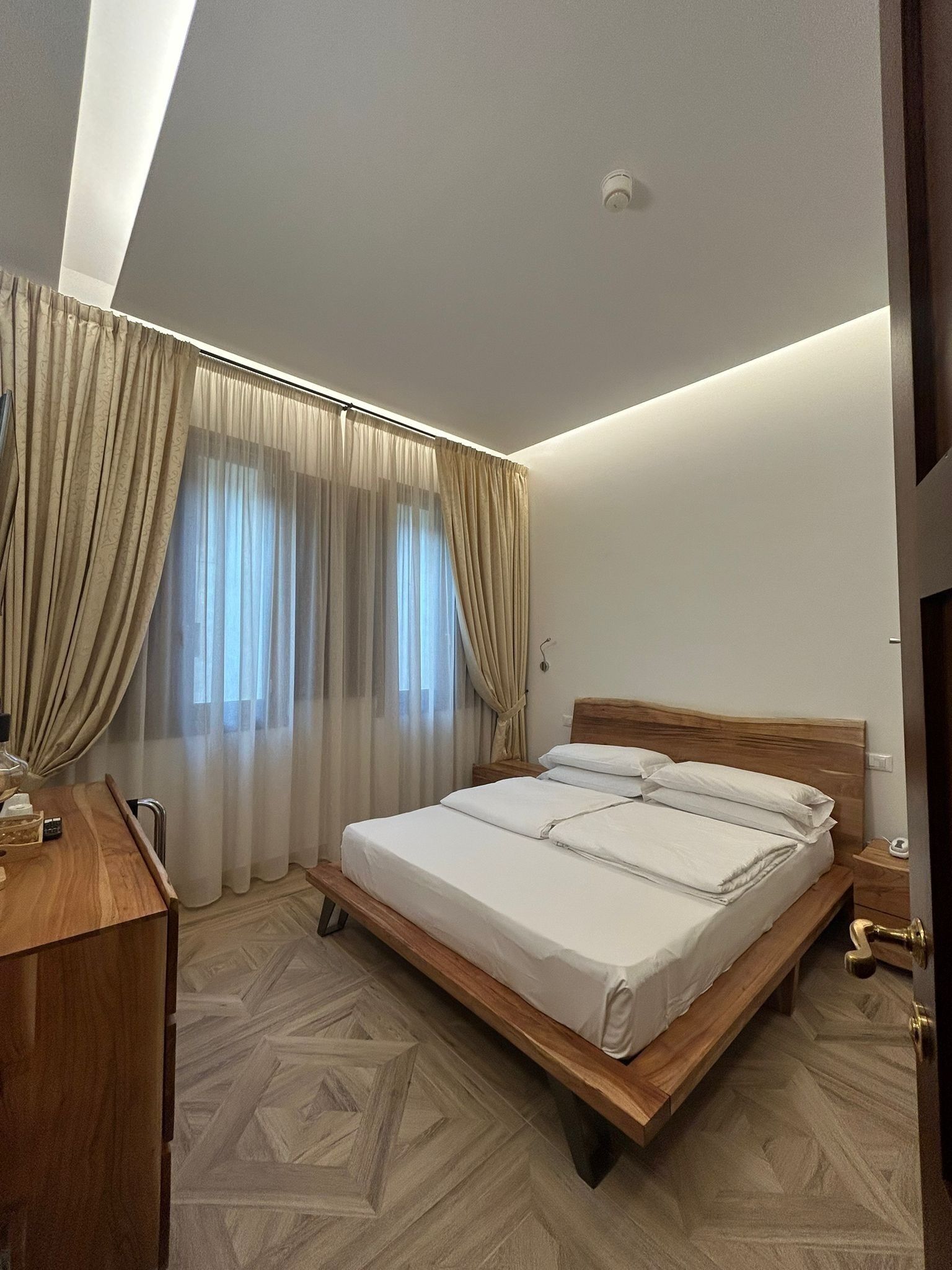 A modern bedroom with a wooden bed, beige curtains, and a geometric patterned floor.