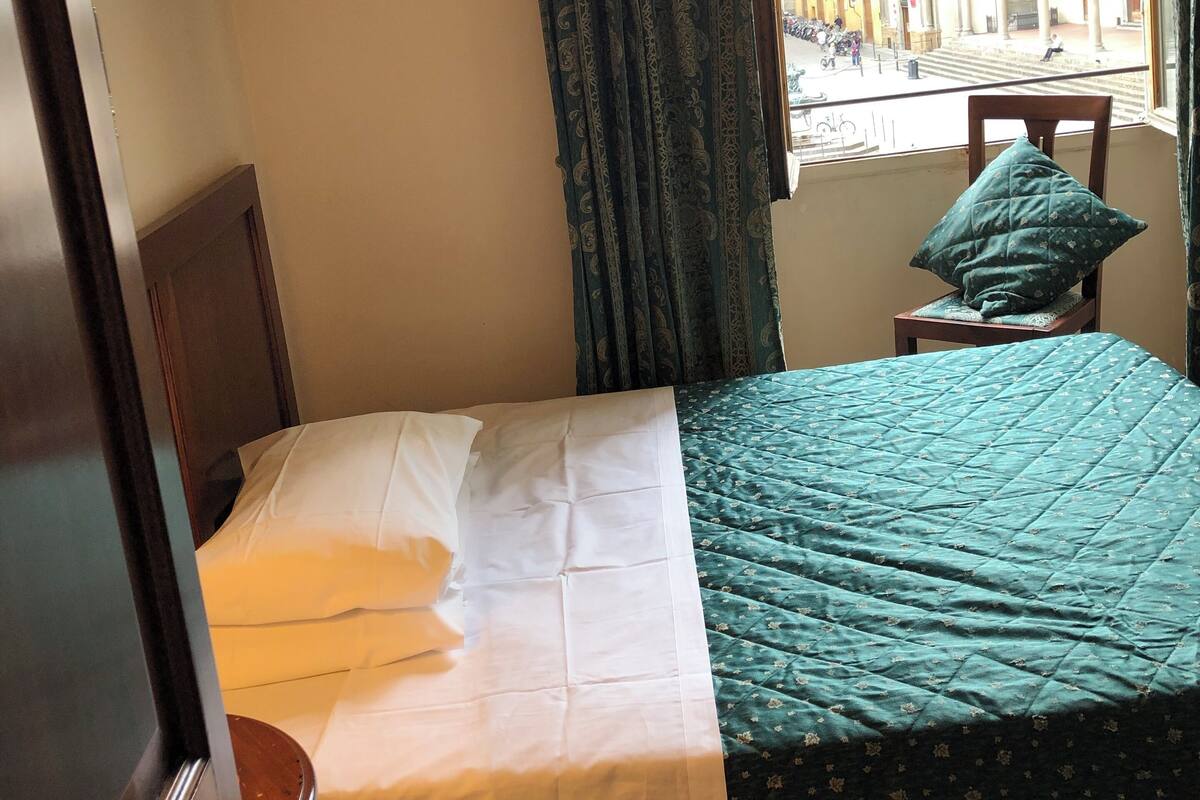 A hotel room with a bed, a chair, a window with curtains, and a view of a building with arches.