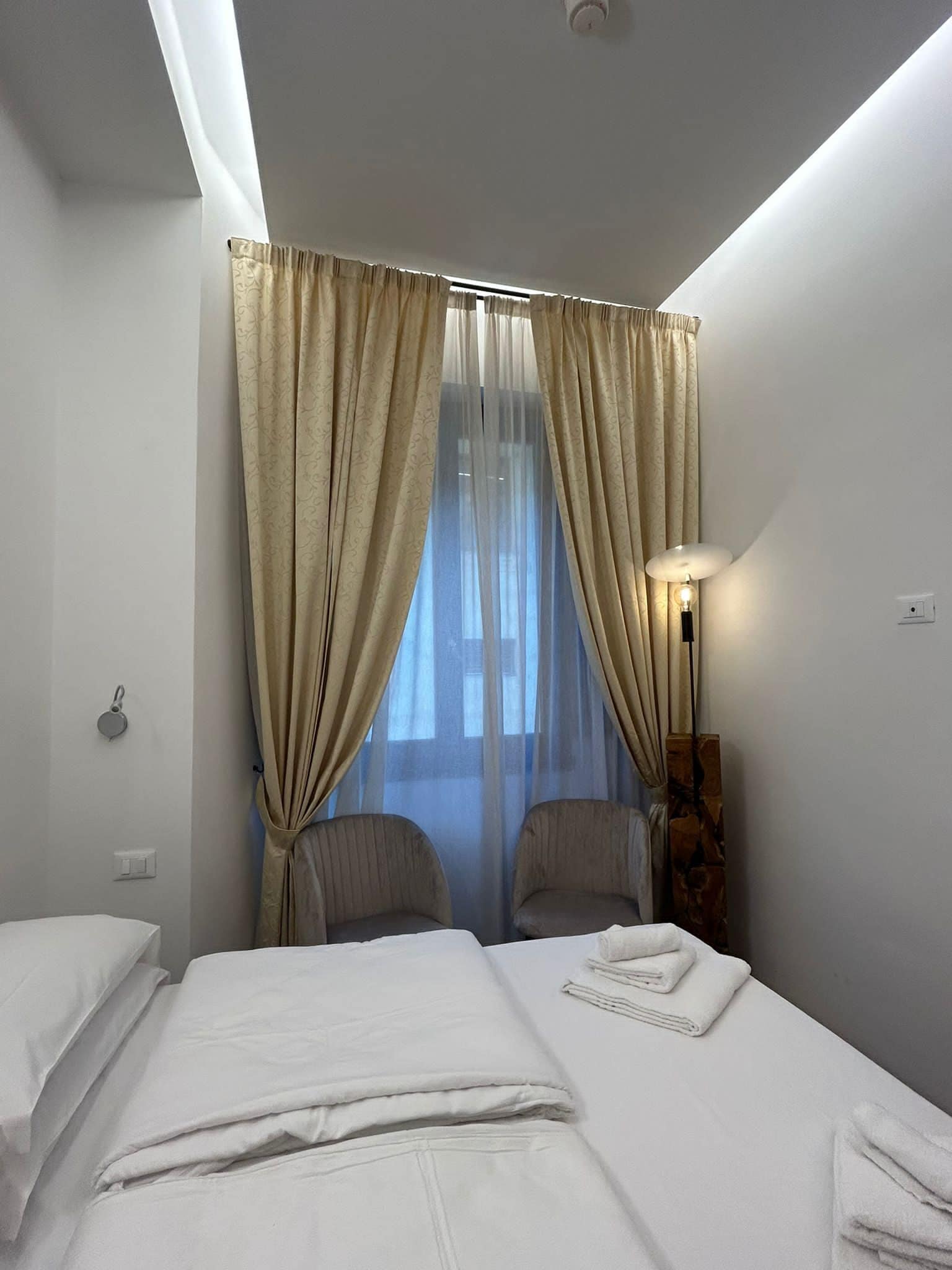 A hotel room with two beds, a nightstand, a lamp, and a window with curtains.