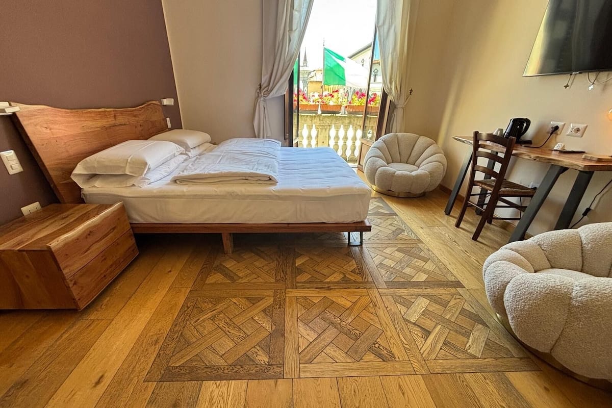 A bedroom with a wooden bed, a small wooden chest, a round armchair, a desk with a chair, and a TV on the wall.