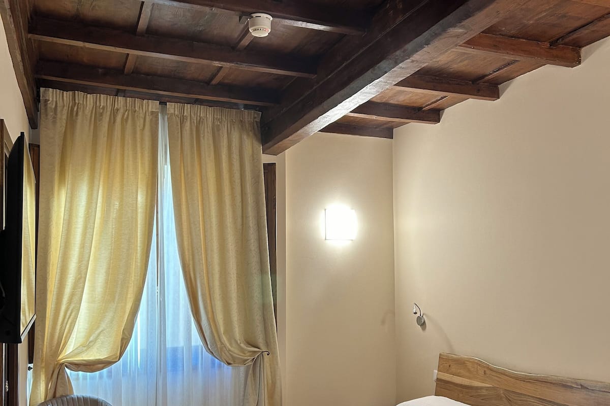 A hotel room with a wooden bed, a chair, a bedside table, and a window with curtains.