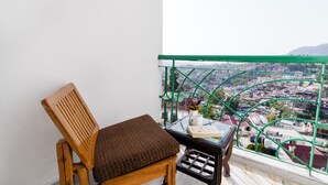 A wooden chair with a cushion, a small table with a book and a vase, and a view of a cityscape through a green railing. - Treebo Anand The Mall Solan (Solan)