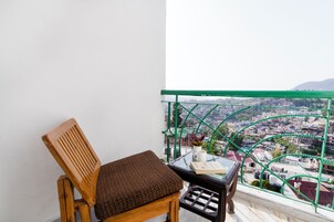 A wooden chair with a cushion, a small table with a book and a vase, and a view of a cityscape through a green railing.