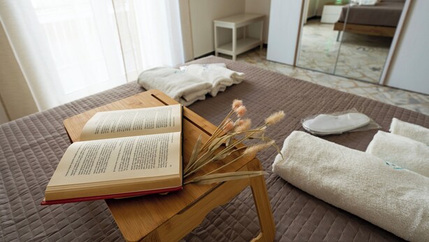 Una cama bien hecha con un libro y flores secas sobre una mesita de madera.
