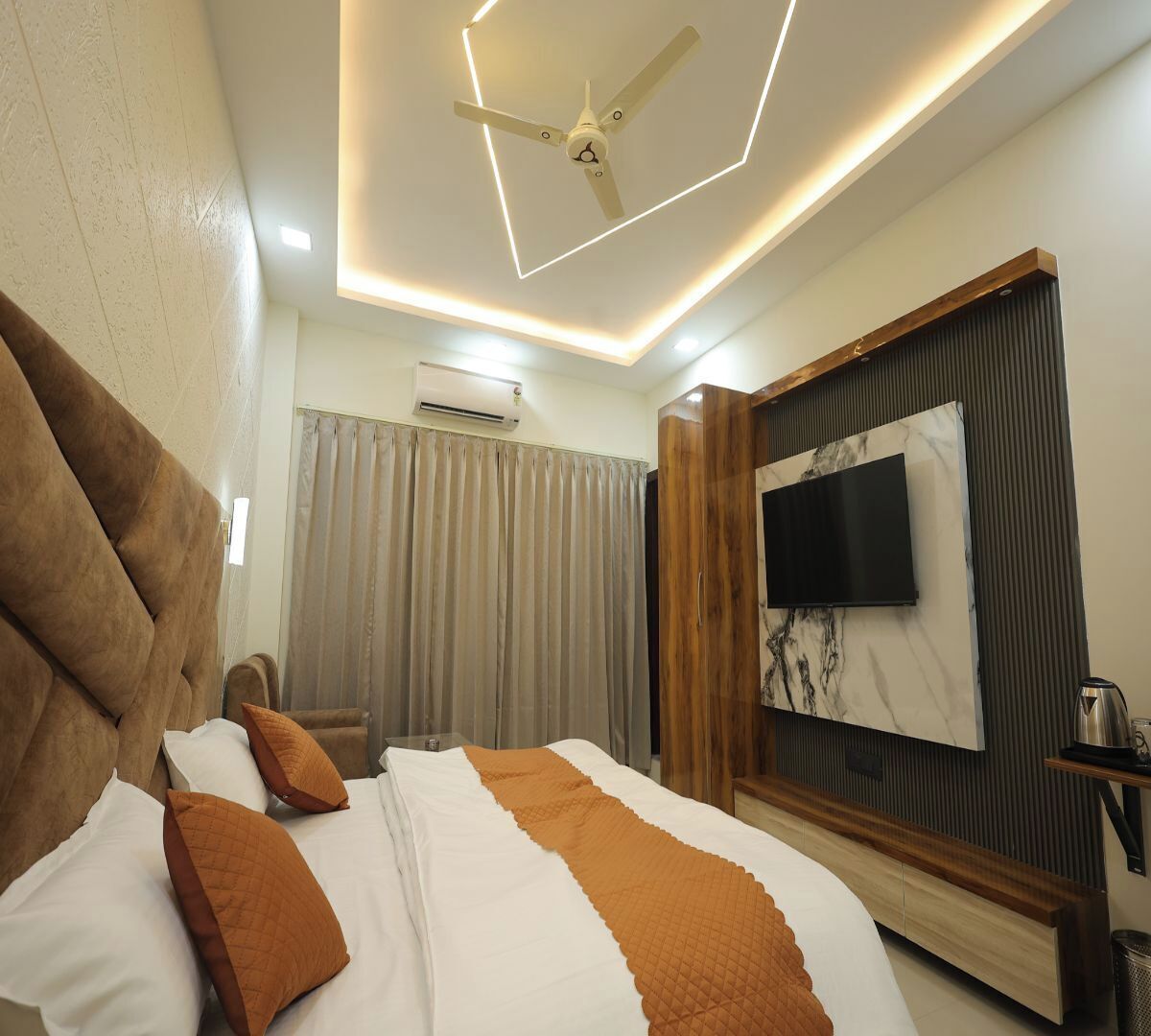 A modern hotel room with a large bed, a ceiling fan, a flat-screen TV mounted on a wooden panel, and a coffee maker on a side table.