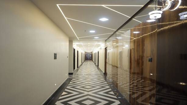 A modern hotel hallway with geometric patterned flooring, recessed lighting, and a glass elevator. - Aayu Residency (Vikasnagar)