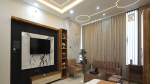 A modern bedroom with a large bed, a TV mounted on a marble wall, a sofa, and a coffee table. - Aayu Residency (Vikasnagar)