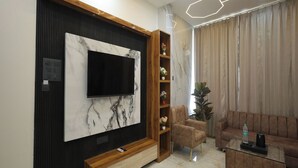 A modern living room with a large flat-screen TV mounted on a marble wall, a brown sofa, a glass coffee table, and a wooden entertainment unit. - Aayu Residency (Vikasnagar)