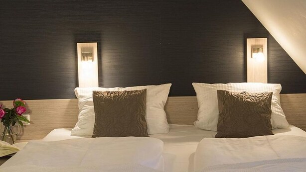 A hotel room with a dark wooden headboard, two bedside lamps, and a bed with white linens and brown pillows.