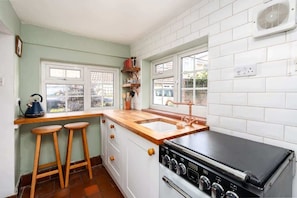 A kitchen with a stove, sink, stools, and a window overlooking a garden.