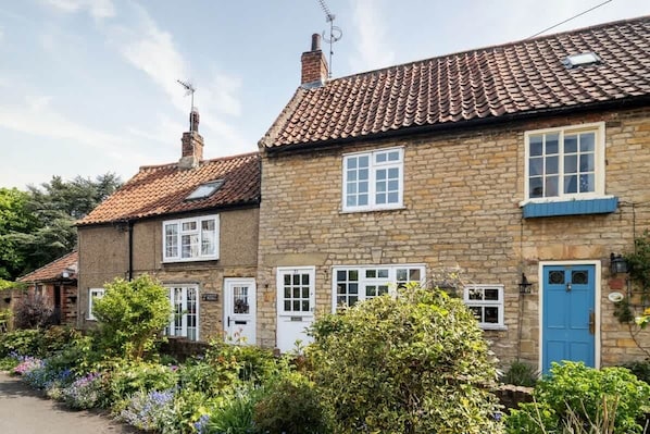 A stone cottage with a tiled roof, a blue door, and a garden with various plants.