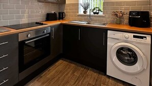 A modern kitchen with a built-in oven, a dishwasher, and a window with vertical blinds. - Wigan 1BR Haven:  Perfect for a Relaxing Stay (Wigan)