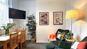 A modern living room with a dining area, a green sofa, a floor lamp, and a TV mounted on the wall. - Wigan 1BR Haven:  Perfect for a Relaxing Stay (Wigan)