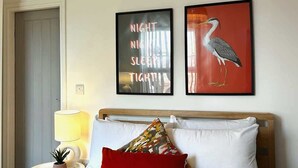 Een net opgemaakt bed met een rode en witte sprei, twee kussens en opgerolde handdoeken. Twee ingelijste foto's hangen aan de muur, één met een vogel en de andere met tekst. Een lamp en een klein plantje staan op een nachtkastje.