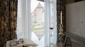 Una habitación con vistas a un castillo, una mesa de cristal con un libro y vasos, una vela y una copa de vino tinto.