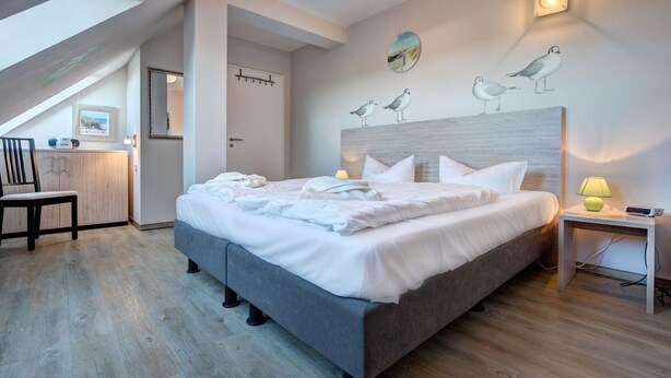 A modern bedroom with a large bed, a desk, and a chair. There is a painting of birds on the wall and a window with a view of the sky.
