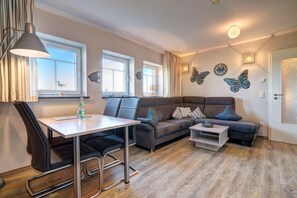 A modern living room with a sofa, a dining table, and a coffee table. There are three windows providing natural light, and decorative wall art is visible. - Windflüchter LH-305 - The country farm Windflüchter LH-305 - Family-friendly (Stolpe auf Usedom)
