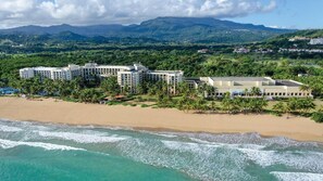 Un hôtel en bord de mer composé de plusieurs bâtiments, doté d’une plage de sable et offrant une vue sur les montagnes en arrière-plan.