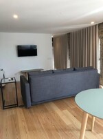 Una sala de estar moderna con un sofá gris, un televisor de pantalla plana montado en la pared, una mesita auxiliar y una mesa redonda con patas de madera.
