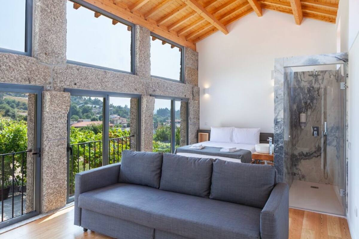 Una habitación moderna con un sofá grande de color gris, una cama con ropa de cama blanca y una ducha con cerramiento de vidrio.