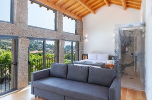 Una habitación moderna con un sofá grande de color gris, una cama con ropa de cama blanca y una ducha con cerramiento de vidrio.